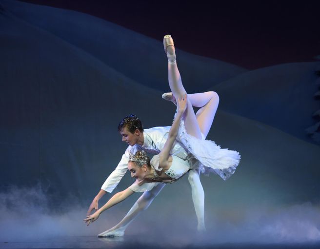 Two ballet dancers perform on stage; the female dancer is en pointe with one leg extended high, supported by the male dancer. Both wear white costumes, and mist covers the stage floor.