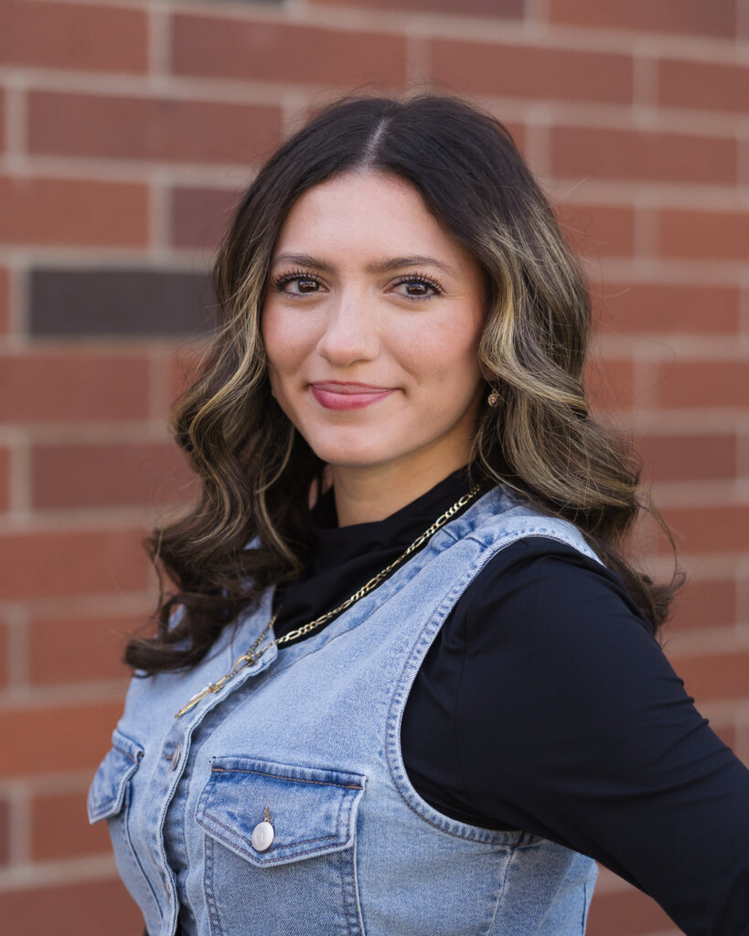 A woman with wavy brown hair and blonde highlights, wearing a denim vest over a black long-sleeve top and a gold chain necklace, stands in front of a brick wall, smiling softly at the camera.