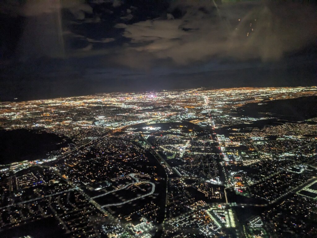 Aerial view of a city at night with streets and buildings illuminated by thousands of lights, stretching to the horizon under a partly cloudy sky.