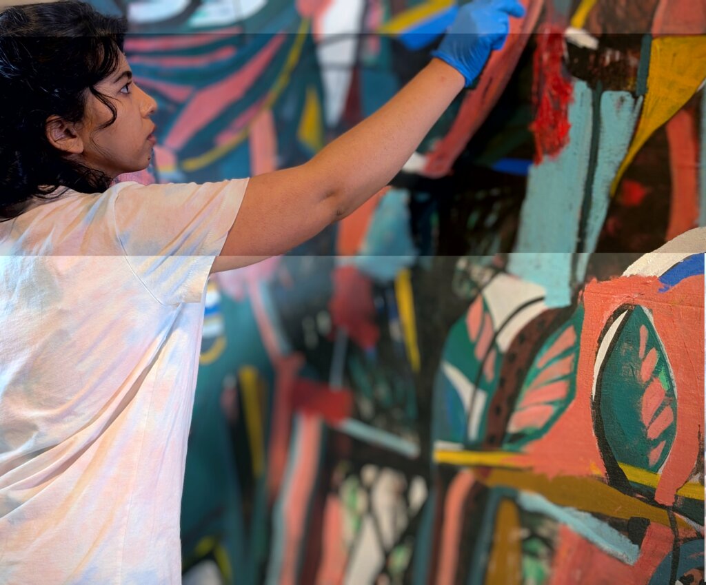 A woman wearing a white shirt and blue gloves paints a colorful, abstract mural on a wall, focusing intently on her work. The mural features bold shapes and vibrant colors.