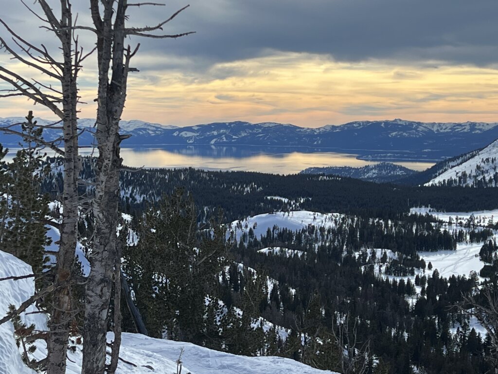 Snow-covered mountains overlooking a serene lake at sunset, with a partly cloudy sky reflecting warm hues. Trees dot the landscape, creating a peaceful vista.
