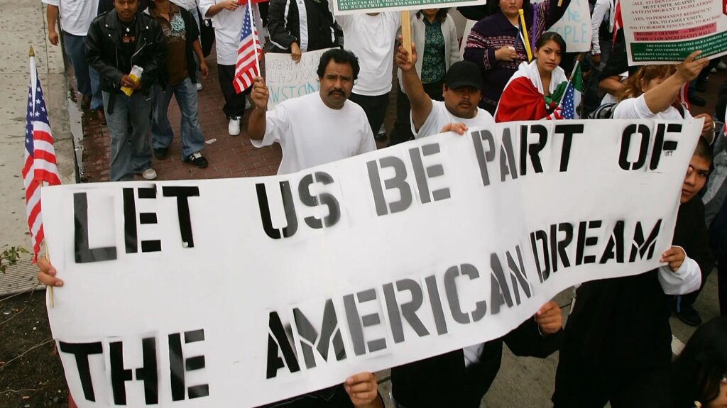 A group of people participate in a protest, holding a large sign that reads “LET US BE PART OF THE AMERICAN DREAM.” Some hold U.S. flags and wear white shirts, and the crowd appears diverse and passionate.