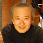 A middle-aged person with short gray hair and a mustache, wearing a dark shirt, sits indoors with wooden pews and brick walls in the background.