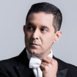A man in a black tuxedo and white bow tie adjusts his collar while looking to the side, against a plain gray background.