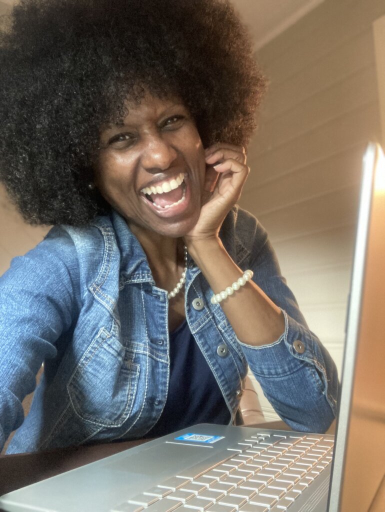 A woman with curly hair and a big smile sits at a table, wearing a denim jacket and pearl bracelet, with her hand on her face and a laptop in front of her. Warm light illuminates her joyful expression.