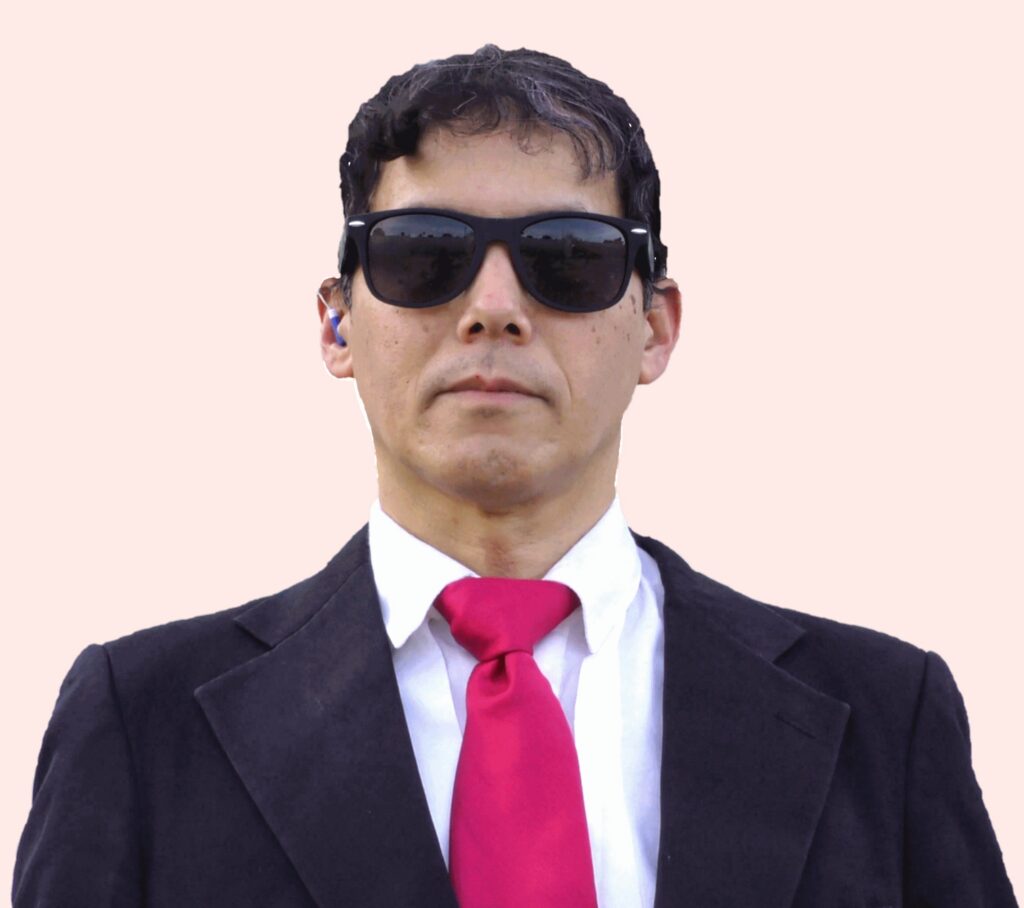 A man wearing a black suit, white shirt, bright red tie, and dark sunglasses stands against a plain light background, looking straight ahead.