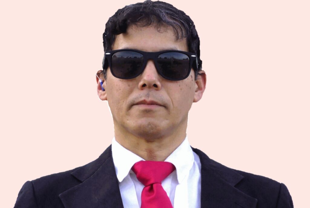A man with short dark hair wears sunglasses, a black suit jacket, white shirt, and a red tie, standing against a plain light background.