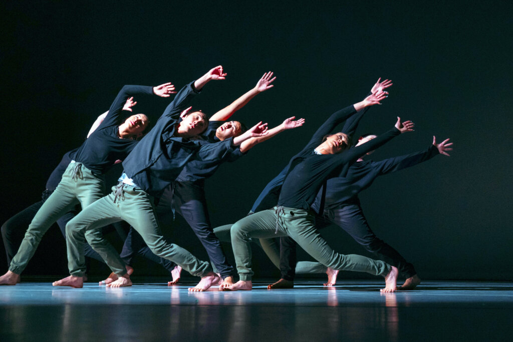 5 dancers in a spotlight on a dark background wearing green pants and black shirts, all leaned back with their arms stretched in the air