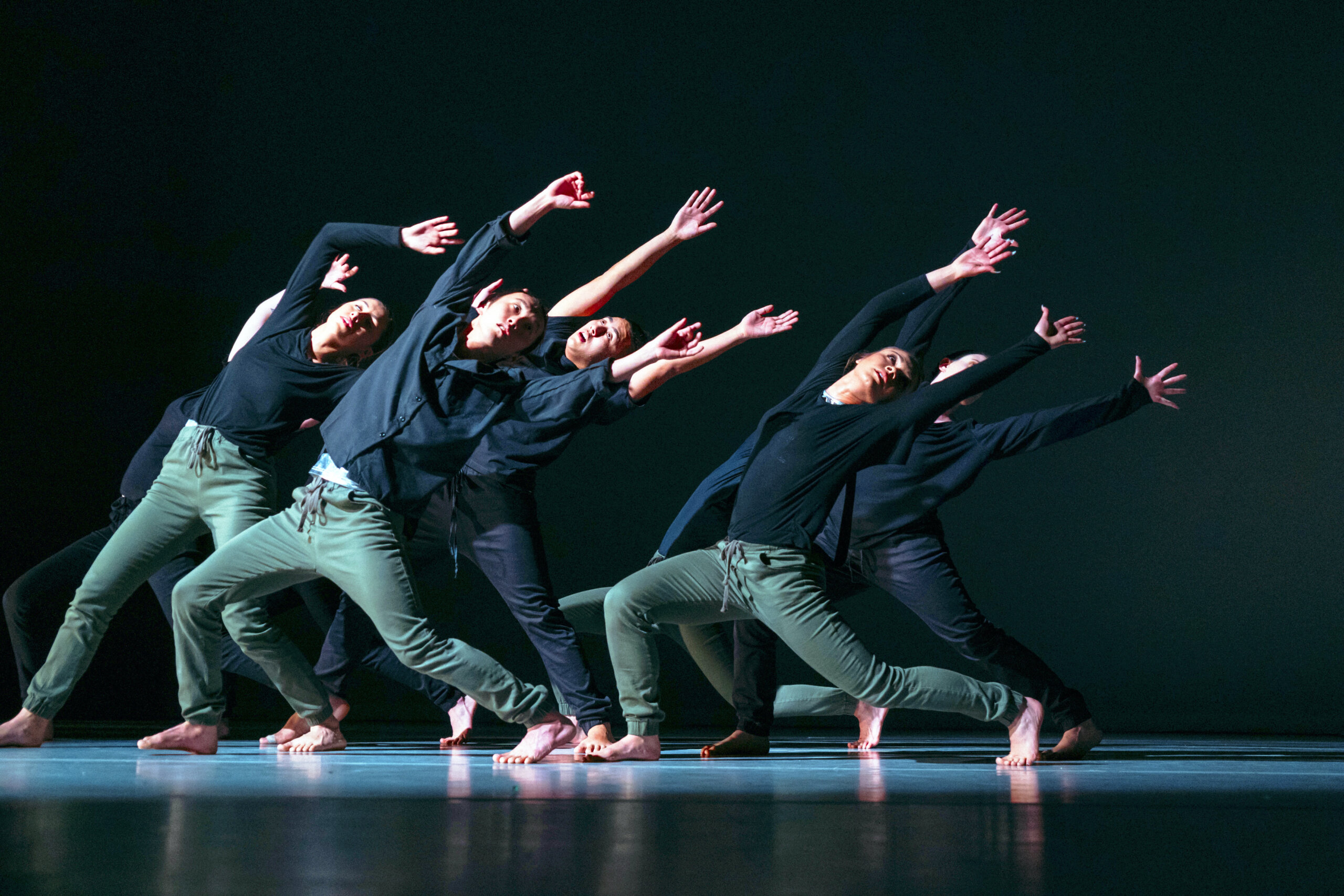 5 dancers in a spotlight on a dark background wearing green pants and black shirts, all leaned back with their arms stretched in the air