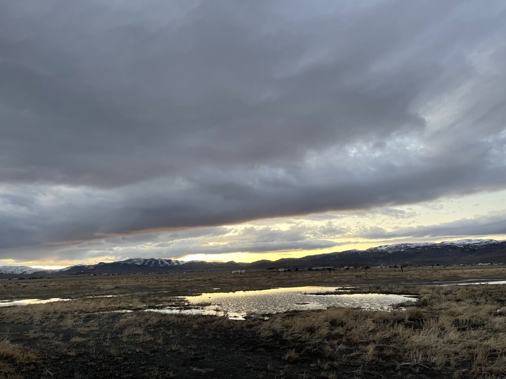 A wide, open landscape with dry grass, puddles of water, and distant snow-capped mountains under a dramatic, cloudy sky at sunset.