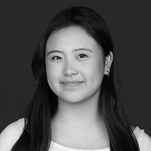 Black and white portrait of a young woman with long dark hair, smiling softly. She is wearing a light-colored top and small hoop earrings, posed against a plain dark background.