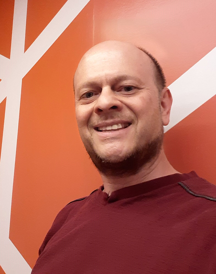 A smiling man with short hair and a beard is wearing a maroon shirt, standing in front of an orange wall with white geometric lines.