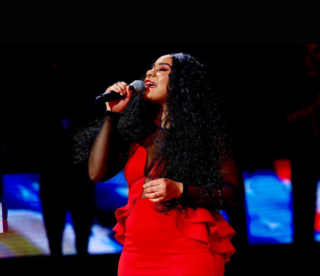 Brittany Soul: A woman in a bright red dress sings passionately into a microphone on stage, with colorful lights and a dark background behind her.