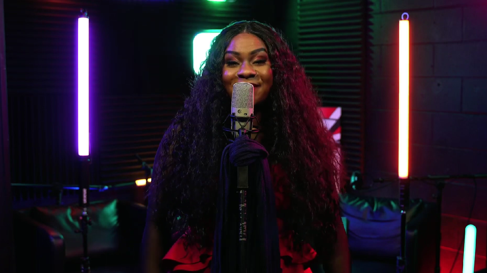 A person with long, curly hair sings into a microphone in a dimly lit studio with colorful neon lights and soundproof foam panels in the background.