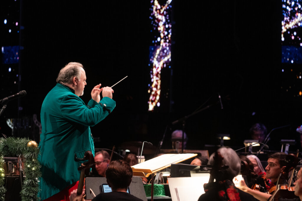 A conductor dress in a green jacket leads an orchestra.