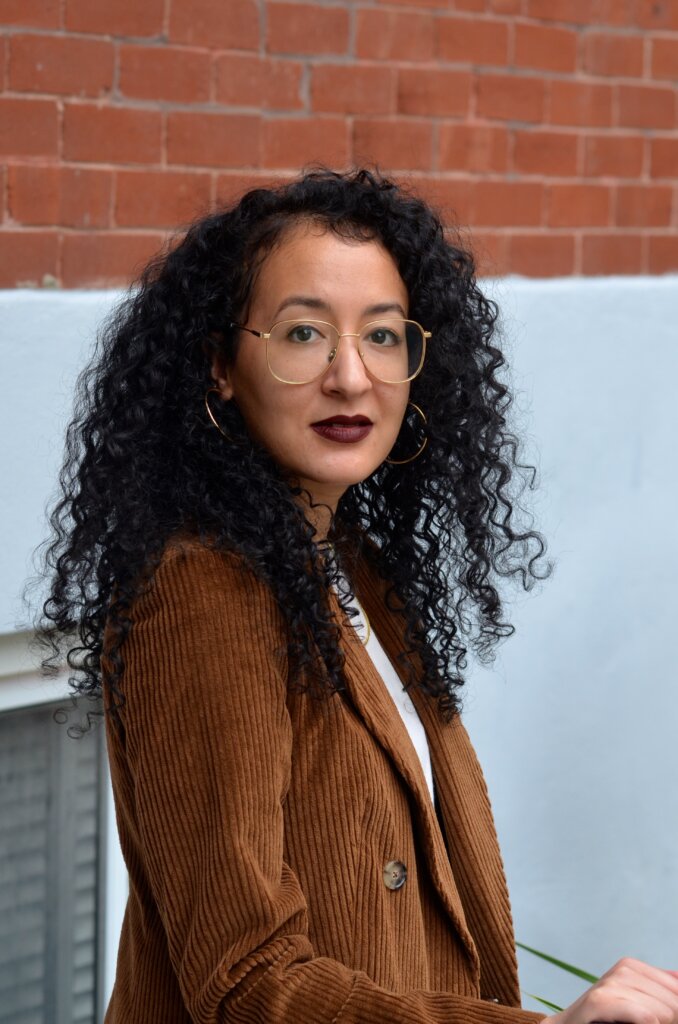 A woman with long curly black hair, wearing round glasses and a brown corduroy blazer, stands in front of a brick and light blue wall, looking at the camera with a neutral expression.