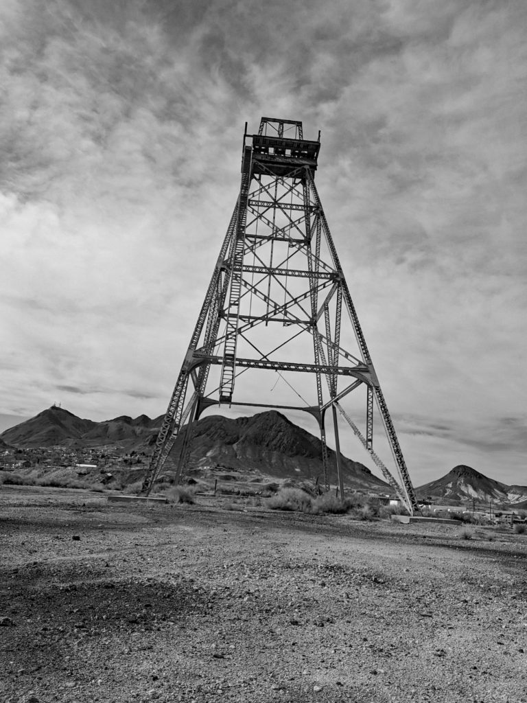 A tall, metal mining tower stands on barren ground with rugged mountains in the background under a cloudy sky; the image is in black and white.
