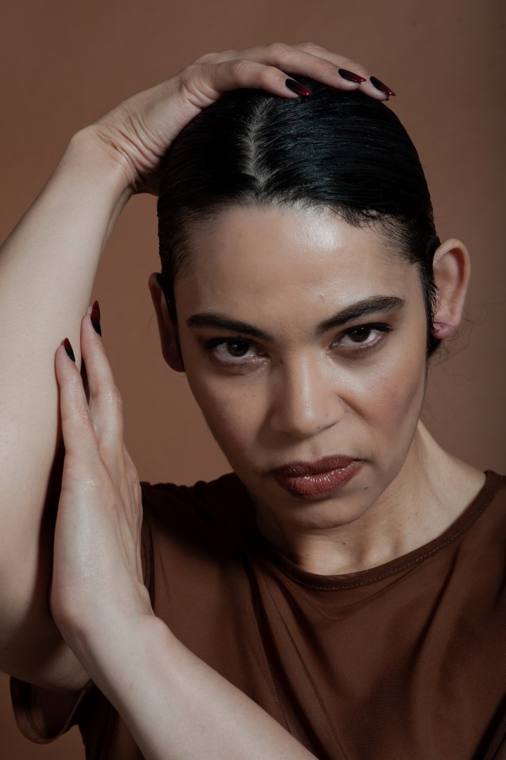 Tonya Todd looks directly into the camera. She has slicked-back dark hair and wears a brown top. One hand is pressed to the side of her head while the other rests on top. Photo credit Michelle Patrick.