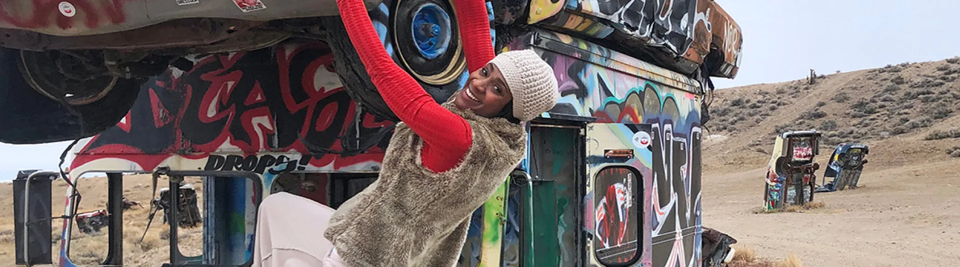 A smiling person in a white hat and furry vest hangs playfully from the wheel of an upside-down, graffiti-covered car stacked on another car in a desert-like outdoor area with more decorated cars in the background.