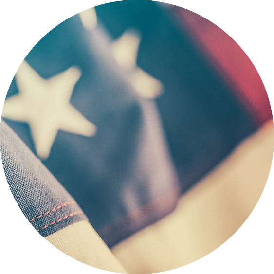 Close-up of a section of the American flag showing white stars on a blue background, with part of the red and white stripes visible, depicted within a circular frame.