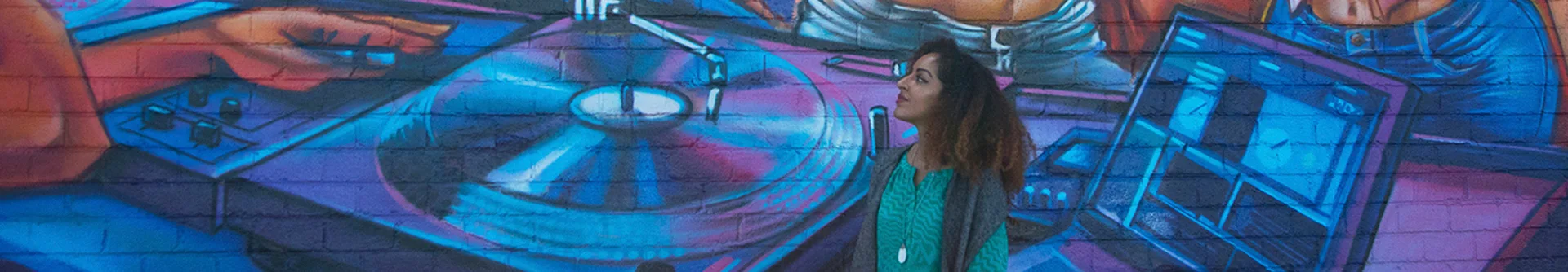 a woman in a teal shirt stands in front of a vibrant mural depicting a dj turntable and hands