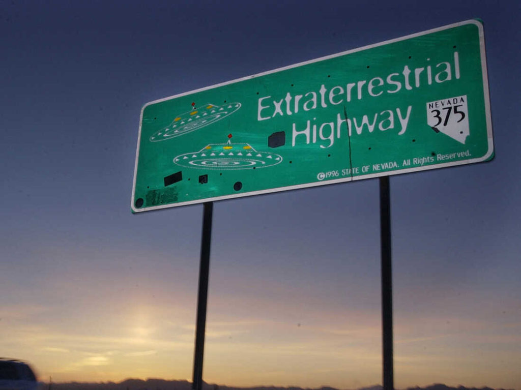 A green road sign at sunset reads “Extraterrestrial Highway” with the Nevada 375 route marker and illustrations of UFOs.