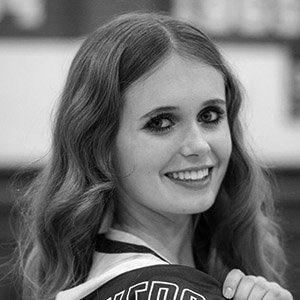 A young woman with long wavy hair smiles at the camera over her shoulder. The photo is black and white, and she appears to be wearing a jersey.