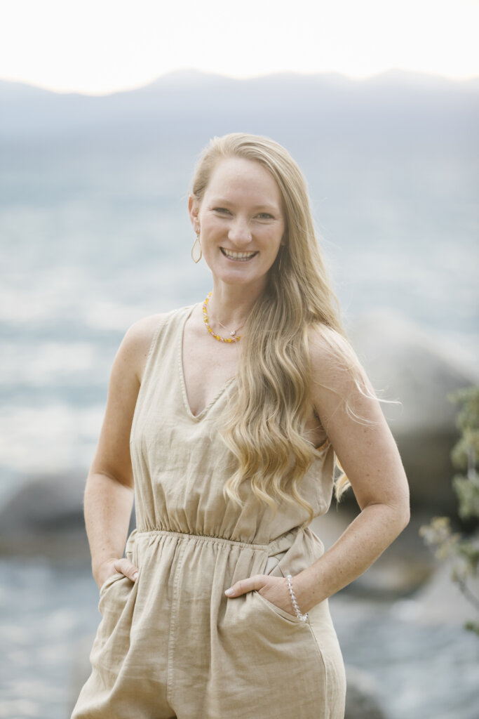 A woman with long, wavy blonde hair stands outdoors by the water, smiling. She wears a sleeveless beige jumpsuit with her hands in the pockets. Large rocks and blurred water are visible in the background.