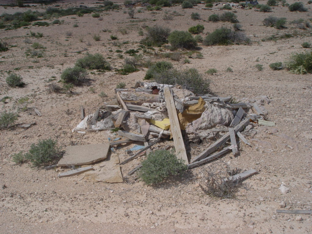 A collapsed wooden structure lies abandoned in a dry, rocky desert landscape, surrounded by scattered bushes and sparse vegetation under a clear sky.