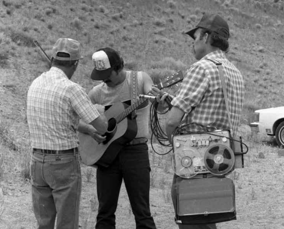 Three men stand outdoors on dry, grassy terrain; one plays guitar, another adjusts the guitar, and the third operates a reel-to-reel tape recorder strapped to his side. A car is partly visible in the background.