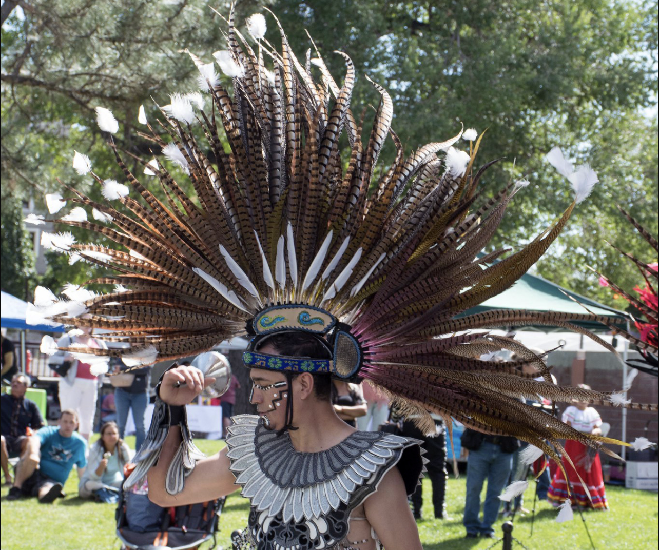 A dancer in traditional Aztec attire with a large feathered headdress performs outdoors, surrounded by people seated on the grass and tents in the background.