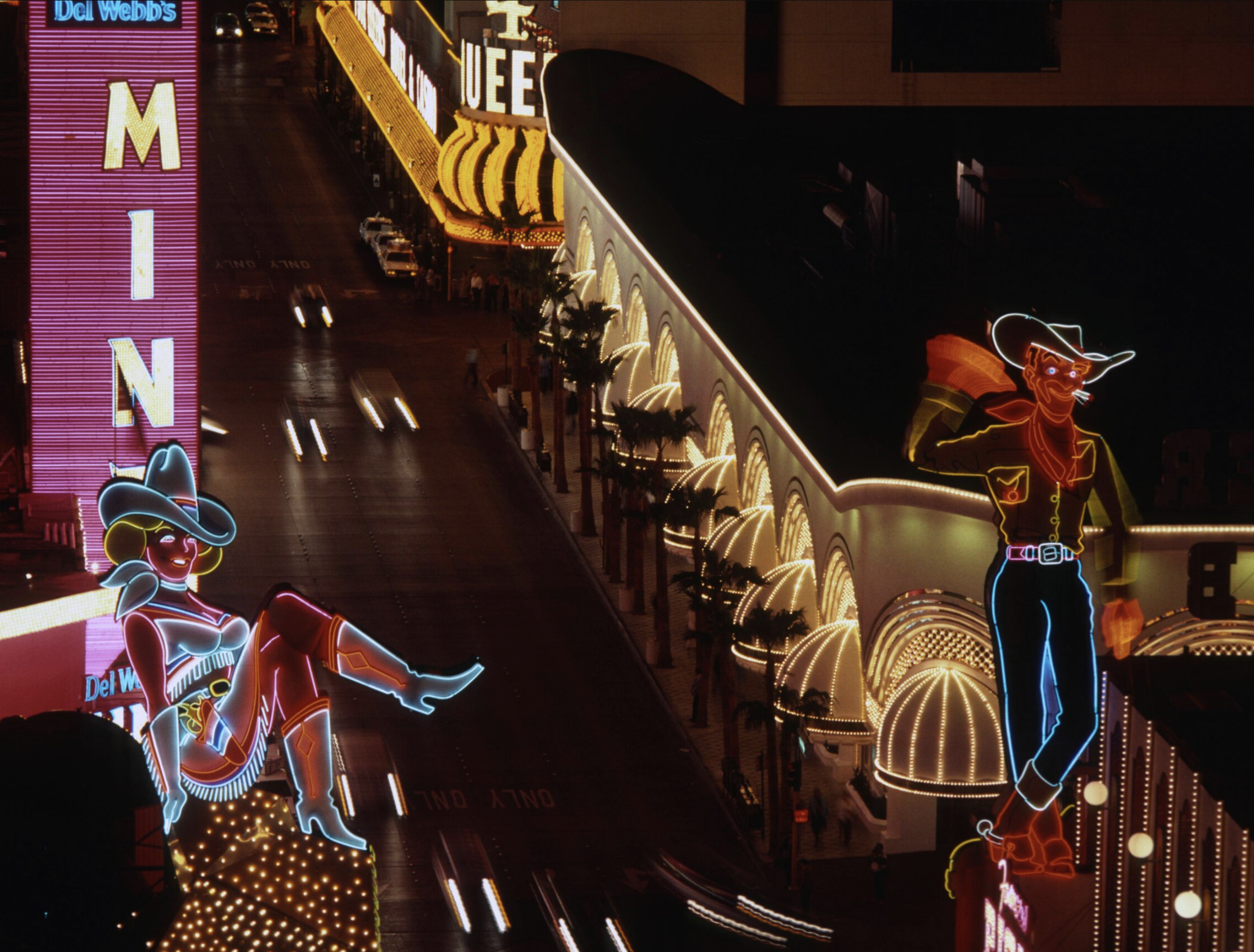 A nighttime street scene in Las Vegas shows bright neon signs, including a large cowgirl sitting and a cowboy standing, surrounded by illuminated buildings and blurred car lights.