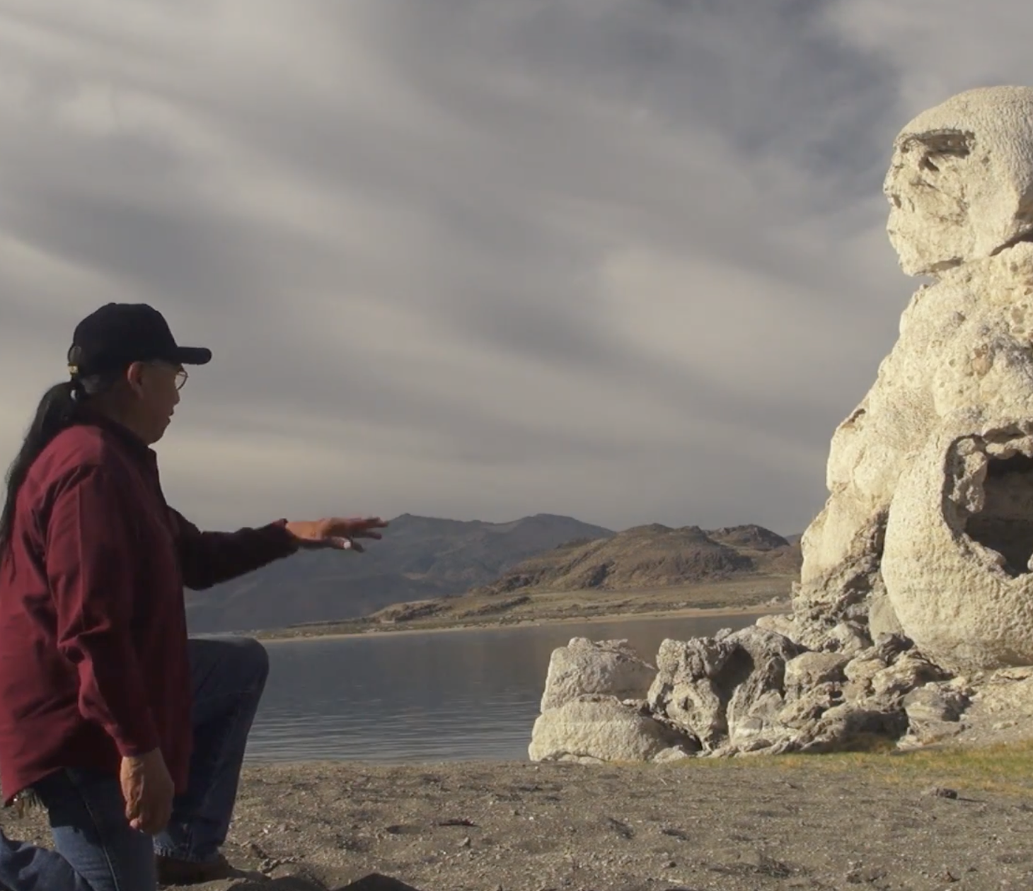 A person in a red shirt and black cap kneels by a lakeshore, facing a large, weathered rock formation that resembles a human face. Mountains and a cloudy sky are in the background.