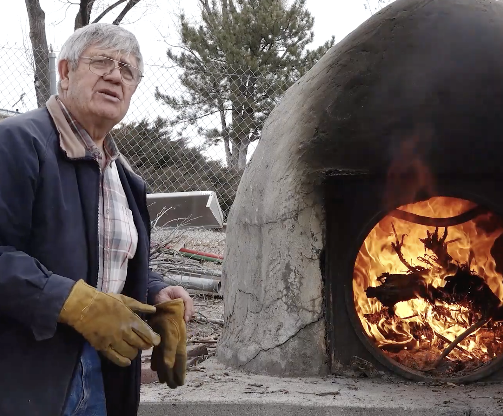 An older man wearing glasses, a jacket, and yellow gloves stands beside a large outdoor wood-fired oven made of stone or clay, with flames and burning logs visible inside. Trees and a fence are in the background.