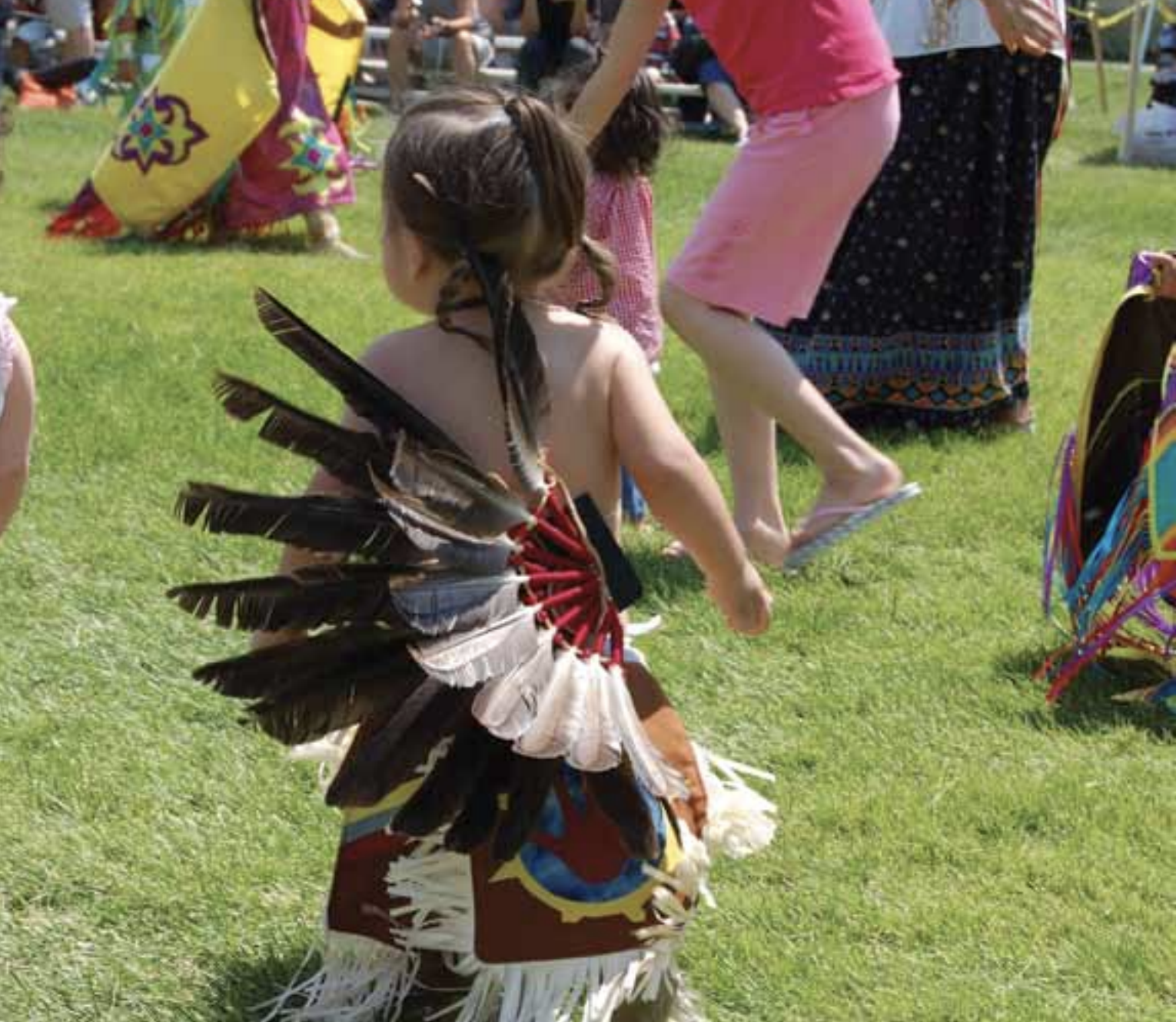 A young child wearing traditional regalia with feathered wings stands on grass among others in colorful clothing, participating in an outdoor cultural event or dance.