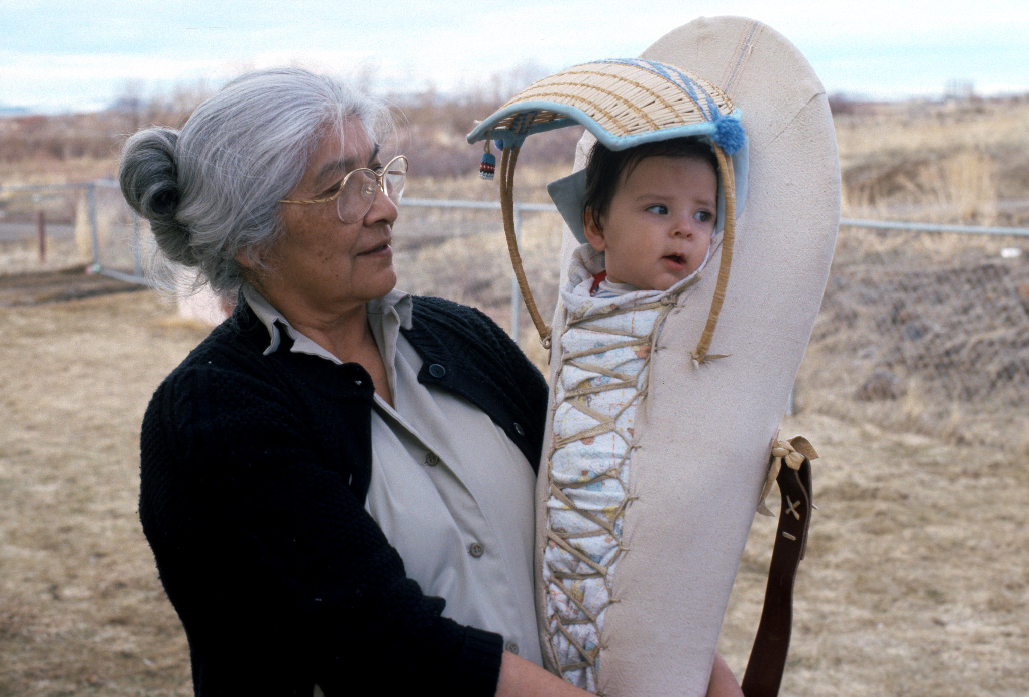 An older woman with gray hair and glasses holds a baby in a traditional Native American cradleboard, outdoors in a field with dry grass and a wire fence in the background.