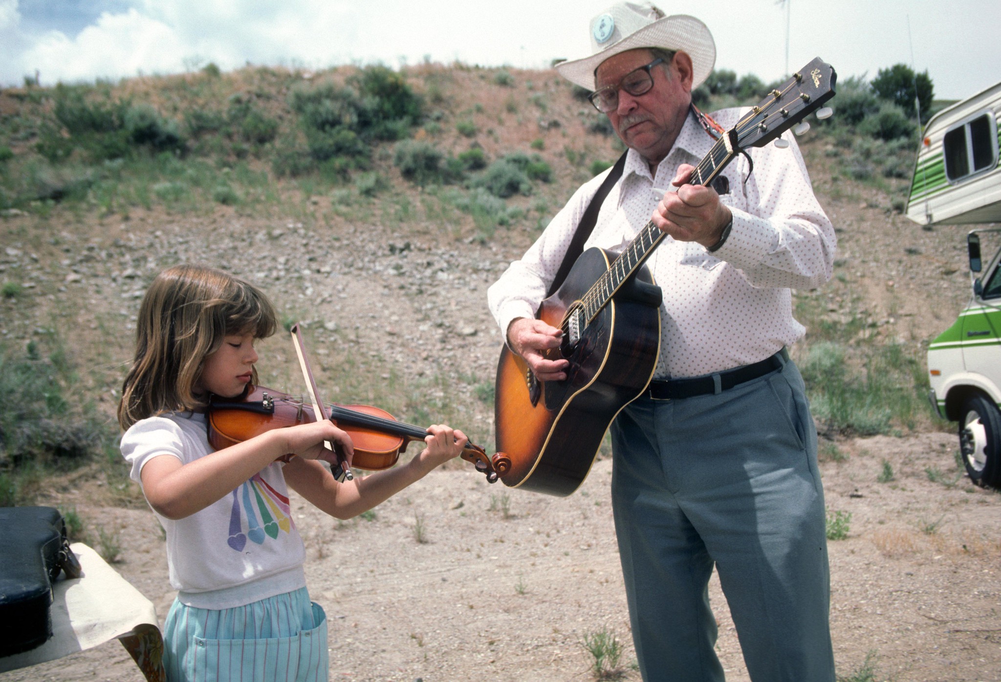 A young girl plays the violin while an older man in a cowboy hat plays an acoustic guitar outdoors, with dry grass and an RV in the background.