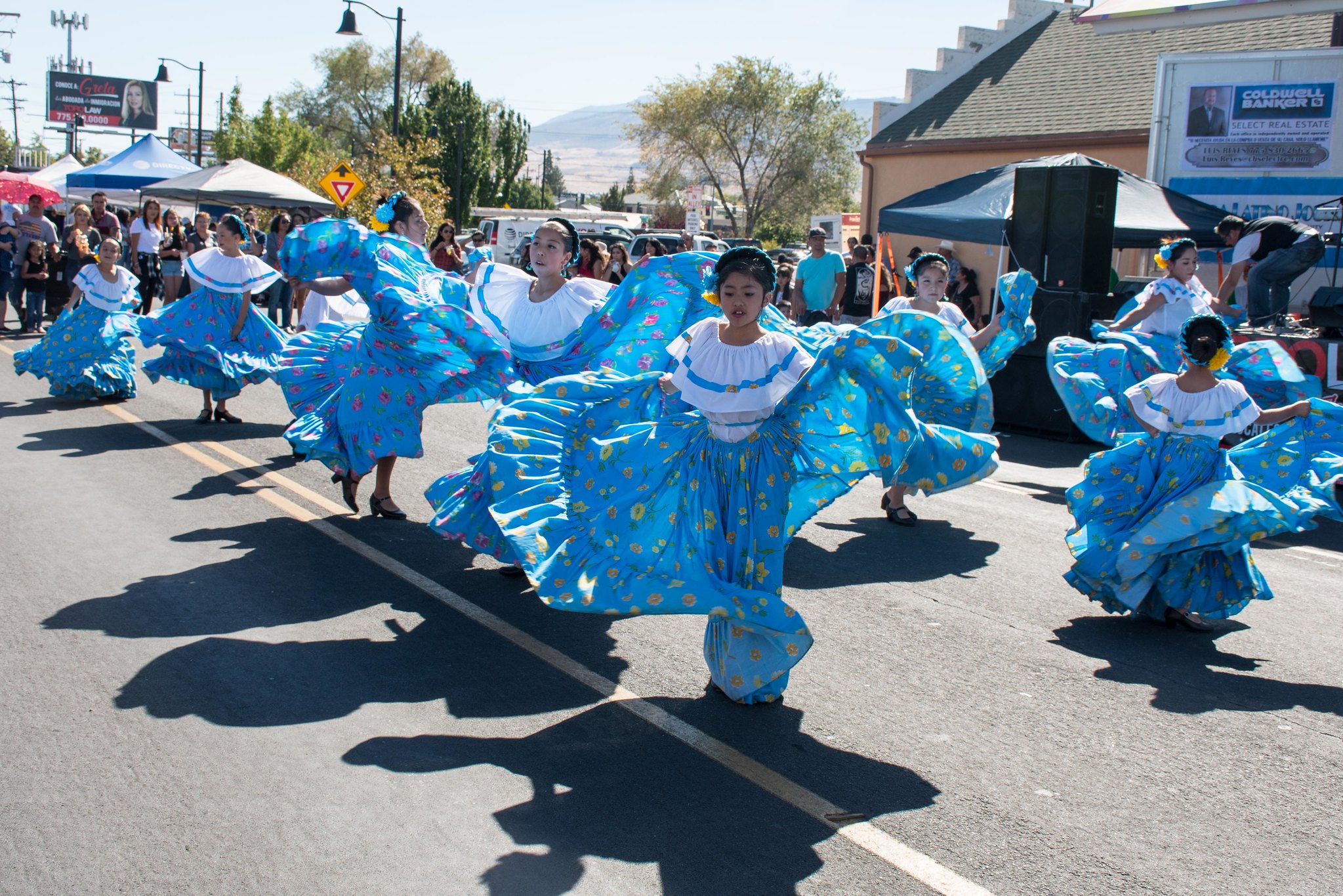 A group of dancers in matching blue, floral skirts and white blouses perform a traditional dance outdoors during a street festival, with onlookers and tents in the background.