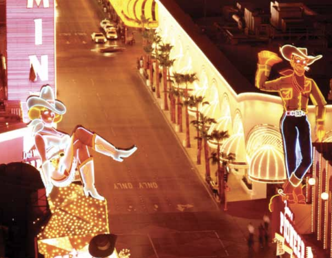 A nighttime street scene features large neon cowboy and cowgirl signs on brightly lit buildings, illuminating an empty road lined with palm trees.