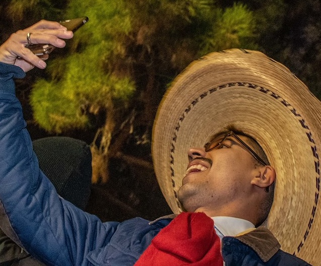 A man wearing a large straw hat, sunglasses, and a blue jacket is smiling and taking a selfie with his phone. He appears to be celebrating outdoors at night, with trees visible in the background.