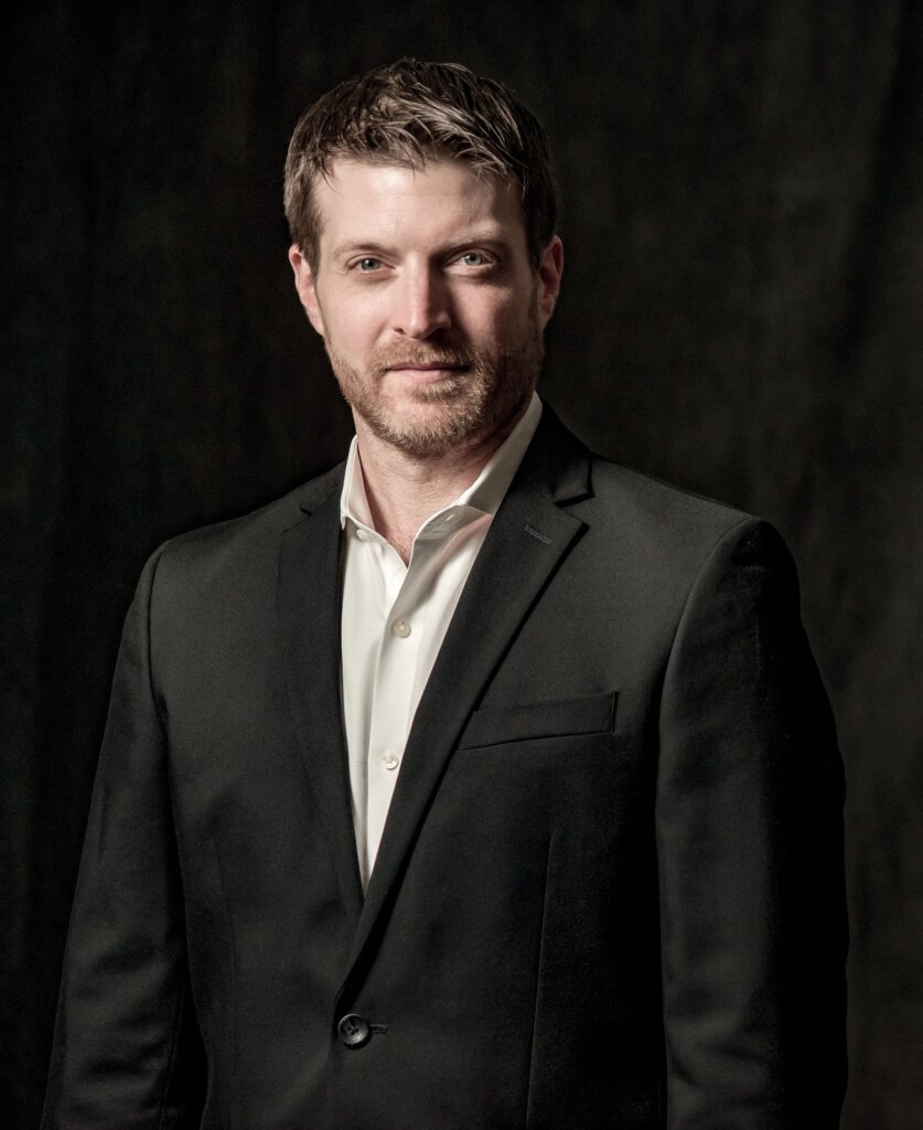 A man with short brown hair and a trimmed beard is wearing a black suit jacket over a white dress shirt, standing against a dark background and looking at the camera with a slight smile.