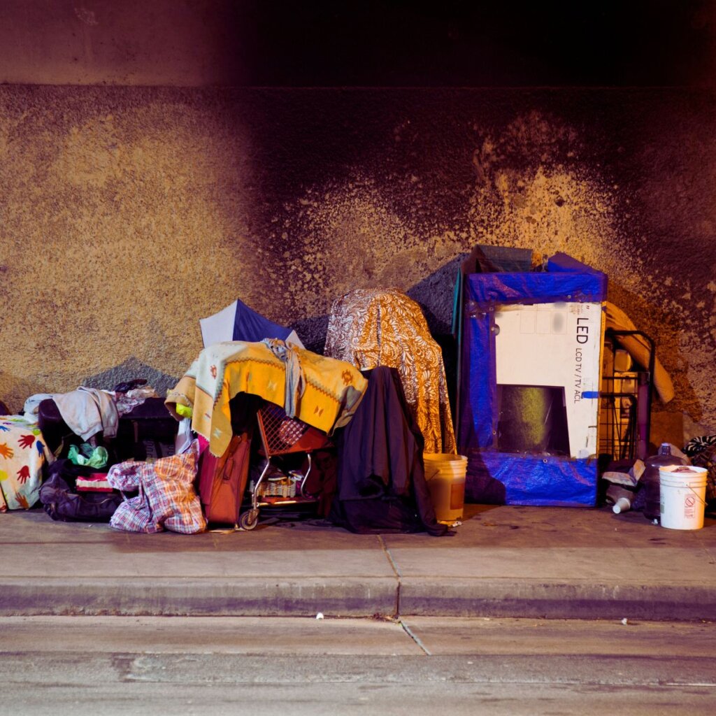 Makeshift shelter on a sidewalk with various belongings, blankets, bags, a plastic-wrapped mirror, and buckets arranged under a wall stained by smoke or dirt.