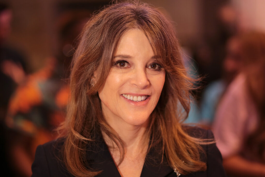 A woman with long, light brown hair and a black top smiles at the camera in a warmly lit indoor setting, with people and colorful blurred backgrounds behind her.