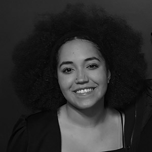 A young woman with curly hair smiles at the camera. She is wearing a dark top and the background is plain and dark.