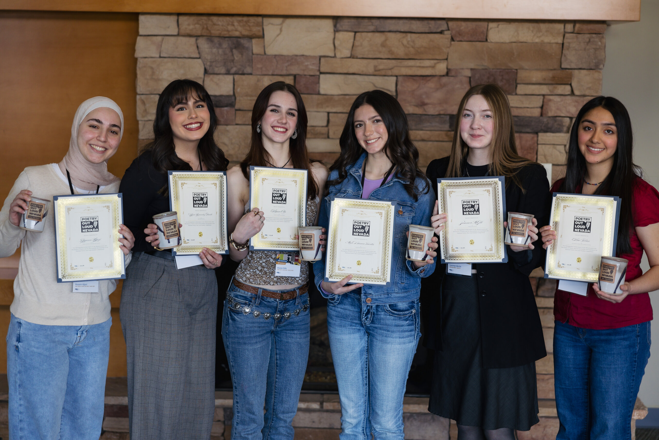 Six young women stand indoors in front of a stone wall, smiling and holding certificates and small boxes, suggesting they have received awards or recognition.