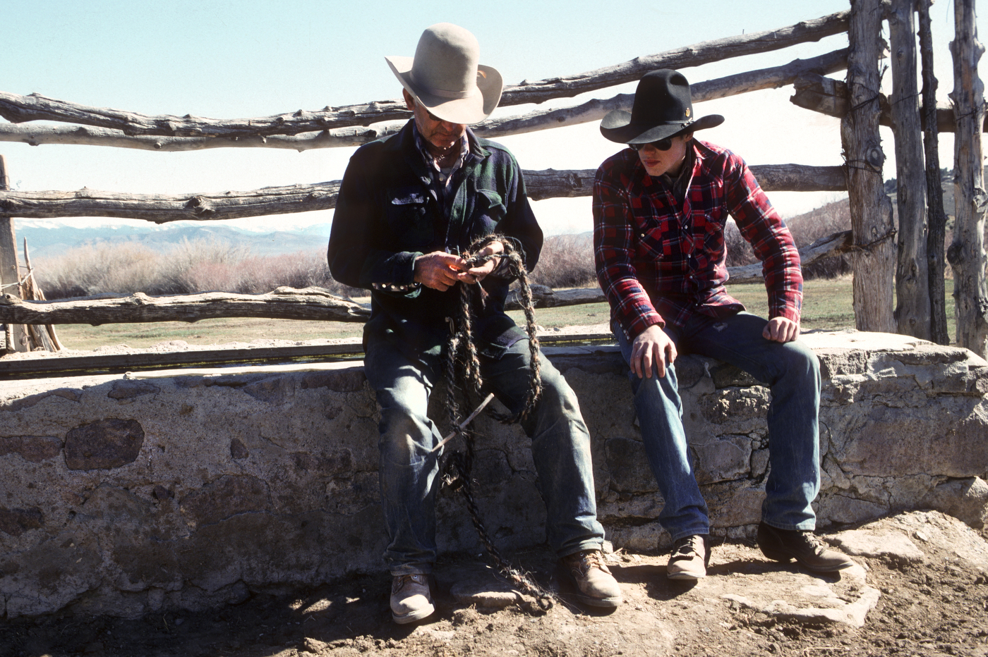 Two people in cowboy hats and casual clothing sit on a stone ledge by a rustic wooden fence in a rural outdoor setting. One inspects a rope or chain, while the other looks on. Mountains and trees are visible in the background.