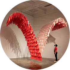 A person stands in a gallery, looking up at a large, V-shaped hanging sculpture made of many red rectangular pieces suspended from the ceiling.